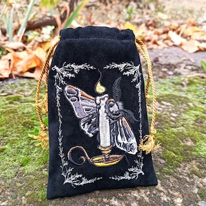 Puede incluir: Bolsa de terciopelo negro con cordón y diseño bordado de polilla y vela. La polilla tiene alas grises y marrones, y la vela tiene una llama amarilla. La bolsa tiene un cierre de cordón dorado y detalles bordados en blanco.