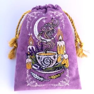 Puede incluir: Una bolsa morada de terciopelo con cordón y un diseño bordado. El diseño presenta una luna creciente, velas, una taza de té y plumas. La bolsa tiene un cierre dorado.