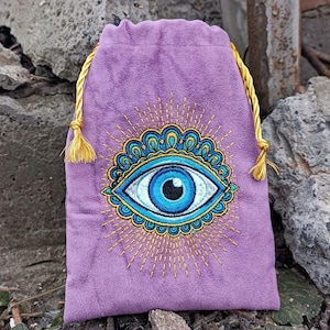 Op de afbeelding: Een paarse fluwelen trekkoordtas met een geborduurd oogontwerp. Het oog is blauw en wit, omgeven door een gouden en blauwgroen decoratief patroon. Gouden koord en kwastjes voor sluiting.