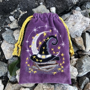 Puede incluir: Una bolsa de terciopelo morado con cordón que presenta un diseño bordado de un sombrero de bruja negro sobre una pila de libros, rodeado por una luna creciente blanca y estrellas amarillas, con cordones amarillos trenzados.