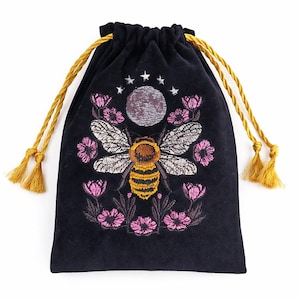 May include: Black velvet drawstring bag with an embroidered bee design. The bee is yellow and brown, surrounded by pink flowers, a moon, and stars. The bag has gold drawstrings with tassels, perfect for storing small items.
