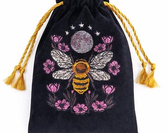 May include: A black drawstring pouch featuring an embroidered design of a central bee with yellow and brown stripes, surrounded by pink flowers and a grey moon topped with five small white stars. The bag has two golden yellow twisted cord ties.
