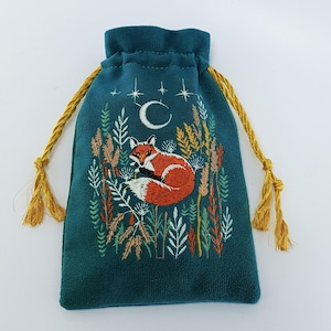 May include: A teal drawstring pouch featuring an embroidered design of a curled-up orange fox surrounded by colorful botanical stalks and grasses. Above the fox, a white crescent moon and small stars are stitched against the fabric surface.