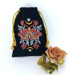 Puede incluir: Una bolsa negra con cordón ajustable que presenta un diseño bordado de una polilla en hilo rojo, amarillo y blanco. El bordado incluye motivos celestiales como estrellas y lunas crecientes, rodeados de elementos botánicos y cordones dorados.