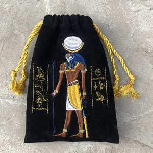 以下が含まれることがあります： 青い頭飾りと白と金の腰布を身に着けたエジプトの神が刺繍された黒い巾着袋。デザインには、頭上の白い太陽円盤、杖、アンク、そして金色のヒエログリフの縦列が含まれています。