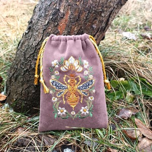 Peut inclure: Un sac mauve à cordon de serrage avec une broderie détaillée d'abeille. L'abeille est dorée avec des détails d'ailes noires et bleues, entourée d'une couronne florale. Le cordon est doré, et le sac est en matière douce et veloutée.