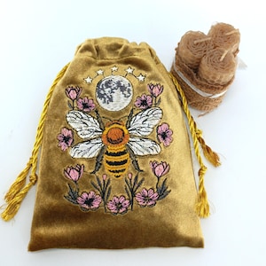 Puede incluir: Una bolsa de terciopelo dorado con cordón que presenta un bordado de una abeja, una luna y flores rosas, acompañada por un conjunto de tres pequeñas velas cilíndricas de cera de abeja marrón claro envueltas en cuerda y una cinta blanca.