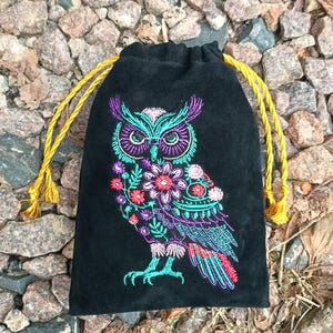 Puede incluir: Bolsa negra con cordón y diseño de búho bordado. El búho está representado con hilo turquesa, morado, rosa y rojo, rodeado de detalles florales. La bolsa tiene cordones y borlas amarillas.