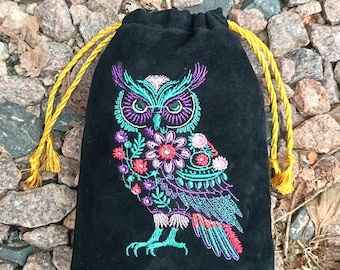 May include: Black drawstring bag featuring an embroidered owl design. The owl is rendered in teal, purple, pink, and red thread, with floral details. The bag has yellow drawstrings and tassels.