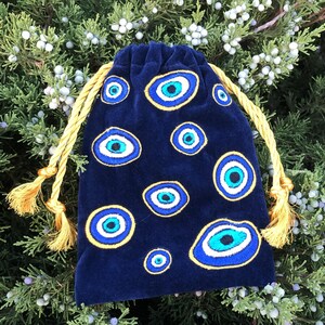 Op de afbeelding: Een donkerblauw fluwelen buideltje met trekkoord, voorzien van meerdere geborduurde boze oog-motieven in tinten blauw, wit en zwart. Het tasje wordt dichtgetrokken met twee dikke, goudgele gedraaide koorden die eindigen in bijpassende kwastjes.