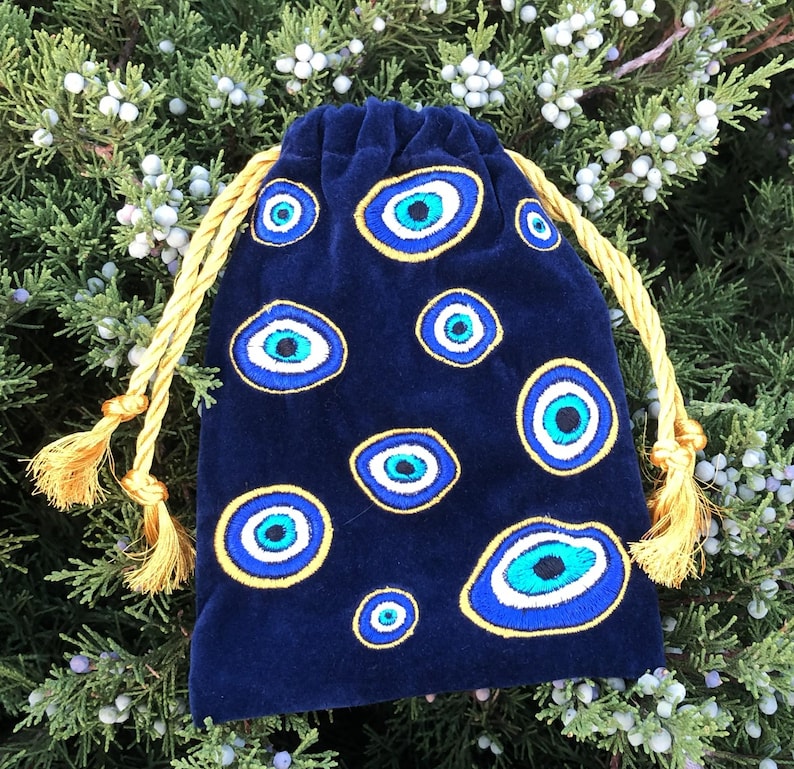 Op de afbeelding: Een donkerblauw fluwelen buideltje met trekkoord, voorzien van meerdere geborduurde boze oog-motieven in tinten blauw, wit en zwart. Het tasje wordt dichtgetrokken met twee dikke, goudgele gedraaide koorden die eindigen in bijpassende kwastjes.