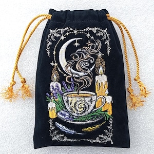 May include: Black drawstring bag with gold cord and tassels. The bag features an embroidered design of a teacup, candles, moon, and stars. The design includes lavender and feathers, creating a mystical theme.