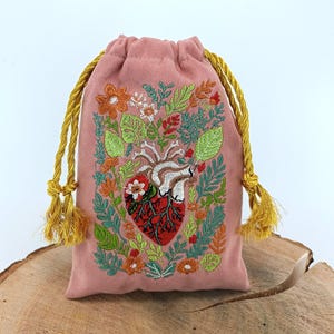 Peut inclure: Une pochette à cordon rose poudré ornée d'un cœur anatomique brodé, entouré de feuillage vert vif et de fleurs orange. Le sac est resserré par deux cordons torsadés en or métallique épais, terminés par des pompons dorés assortis.