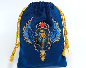 May include: A deep blue velvet drawstring pouch featuring an embroidered winged scarab beetle design in gold, blue, and red thread. The pouch has two gold twisted cord drawstrings at the top, providing a secure closure for the small fabric bag.