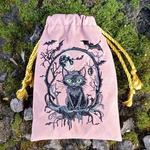 Puede incluir: Una bolsa de terciopelo rosa con cordón y un diseño bordado con un gato negro de ojos verdes, una luna llena, murciélagos y ramas. La bolsa tiene cordones y borlas doradas. El diseño tiene un tema gótico o de Halloween.