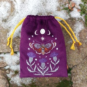 Puede incluir: Una bolsa de color morado oscuro con cordón, decorada con un bordado detallado de una polilla sobre tres flores de cardo. Sobre la polilla, un ciclo de fases lunares y una estrella están bordados en hilo blanco. Cordones de color amarillo.