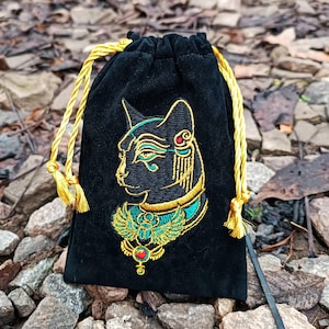 May include: Black velvet drawstring bag with an embroidered Egyptian cat design in gold, teal, and red. The bag has a gold drawstring closure and tassels. The design is reminiscent of ancient Egyptian art and is suitable for storing small items.