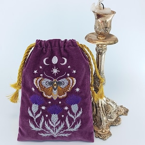 Puede incluir: Una bolsa de terciopelo morado con cordones dorados y bordados detallados de una polilla, fases lunares, estrellas y flores de cardo. Junto a la bolsa hay un candelabro de metal plateado desgastado con una vela parcialmente derretida.