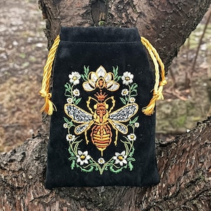 Peut inclure: Pochette noire en velours avec un motif d'abeille brodé. L'abeille est détaillée avec du fil doré, orange et noir, entourée de fleurs blanches et de feuilles vertes. Le cordon de serrage est doré.