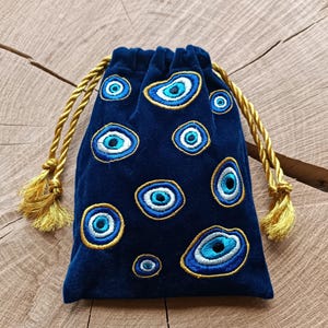Op de afbeelding: Een donkerblauw fluwelen buideltje met trekkoord, voorzien van geborduurde boze oog-motieven in blauwe, witte en zwarte tinten, omlijnd met gouddraad. De tas heeft twee gedraaide gouden koorden met bijpassende kwastjes aan de uiteinden.