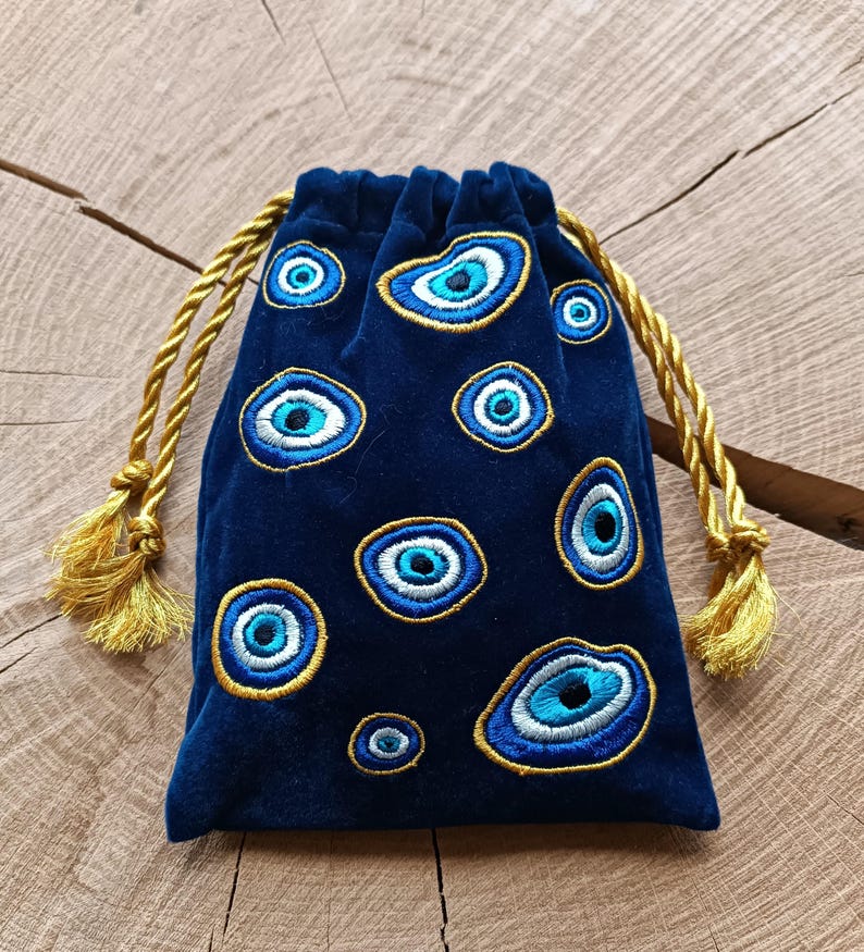 Op de afbeelding: Een donkerblauw fluwelen buideltje met trekkoord, voorzien van geborduurde boze oog-motieven in blauwe, witte en zwarte tinten, omlijnd met gouddraad. De tas heeft twee gedraaide gouden koorden met bijpassende kwastjes aan de uiteinden.