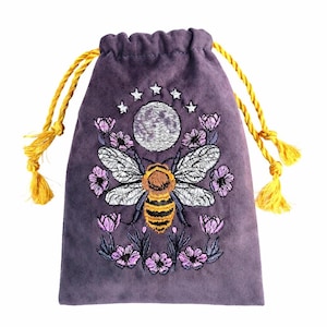 Puede incluir: Una bolsa de cordón morada con un diseño bordado de una abeja bajo una luna y estrellas, rodeada de flores rosas. La bolsa tiene dos cordones trenzados amarillos con borlas en los extremos, lo que permite un cierre seguro y práctico.