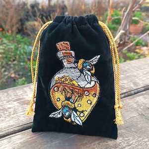 Peut inclure: Sac en velours noir avec cordon doré. Le sac présente une broderie d'une fiole de potion avec deux abeilles. La fiole contient un liquide doré et un bouchon en liège. Les abeilles ont des yeux bleus et des ailes blanches.