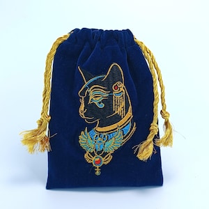 May include: A deep blue velvet drawstring pouch featuring an embroidered black cat head with gold and blue accents, wearing an ornate collar with a winged scarab beetle motif. The bag is cinched with two twisted gold metallic cord drawstrings.