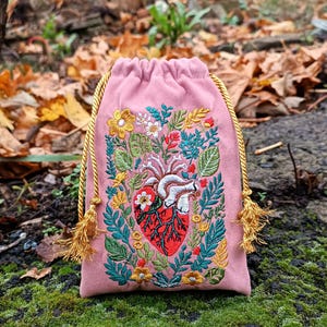 Peut inclure: Un sac rose à cordon de serrage avec un motif de cœur anatomique brodé. Le cœur est rouge et blanc, entouré de feuilles vertes et de fleurs colorées. Le sac a des cordons et des glands dorés.