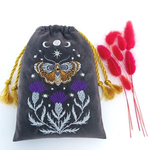 Puede incluir: Una bolsa de terciopelo gris carbón con cordón ajustable, cuerdas trenzadas doradas y bordados intrincados. El diseño incluye una polilla central, fases lunares, estrellas y tres cardos morados con tallos y hojas en tono plateado.
