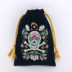 Puede incluir: Una bolsa negra con cordón ajustable que presenta un diseño bordado de una calavera con huesos cruzados, rodeada de enredaderas verdes, flores blancas, detalles en rosa y lunas crecientes doradas. La bolsa tiene dos cordones dorados.