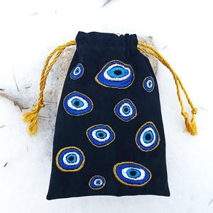 Op de afbeelding: Een zwart trekkoordzakje met geborduurde boze oog-motieven in tinten blauw, wit en goud. De tas wordt gesloten met twee goudgele gedraaide koorden die aan de bovenkant uitsteken, wat zorgt voor een functionele sluiting voor de kleine stoffen tas.
