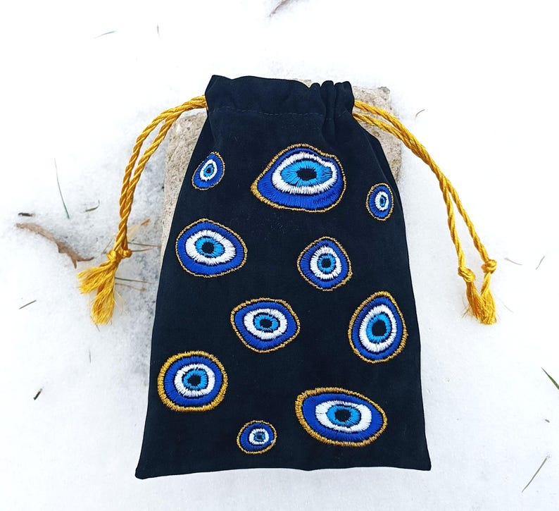 Op de afbeelding: Een zwart trekkoordzakje met geborduurde boze oog-motieven in tinten blauw, wit en goud. De tas wordt gesloten met twee goudgele gedraaide koorden die aan de bovenkant uitsteken, wat zorgt voor een functionele sluiting voor de kleine stoffen tas.