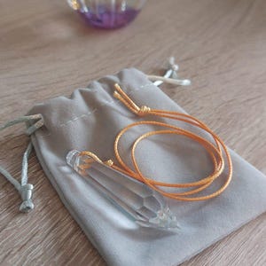May include: A clear crystal pendulum with an orange cord rests on a gray velvet pouch. The pendulum is hexagonal and pointed at the bottom. A glass paperweight with a pink orb is in the background.