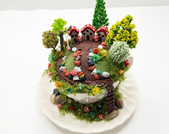 May include: A whimsical miniature fairy garden scene inside a teacup. The scene includes tiny houses with red and white mushroom-shaped roofs, a winding path, colorful stones, and miniature trees. The teacup sits on a white saucer.