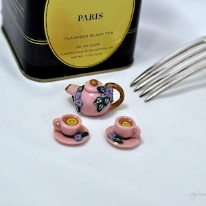 May include: A miniature pink teapot with purple flowers and green leaves, two pink teacups with saucers, and a box of Paris flavored black tea.