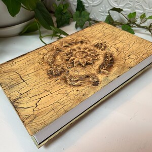 May include: A rectangular book with a distressed, crackled gold finish on the cover. The center of the cover features a raised, carved circular floral motif with intricate petal details. The pages of the book are white and visible along the side.