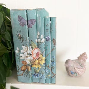 May include: A set of decorative books with a teal background and floral and butterfly designs. The books are arranged vertically, with the spines facing forward. A small, decorative hen figurine is to the right of the books.