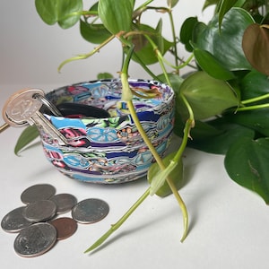May include: A small, colorful bowl with a blue, white, and green floral pattern. The bowl is filled with keys and coins. A green plant is in the background.