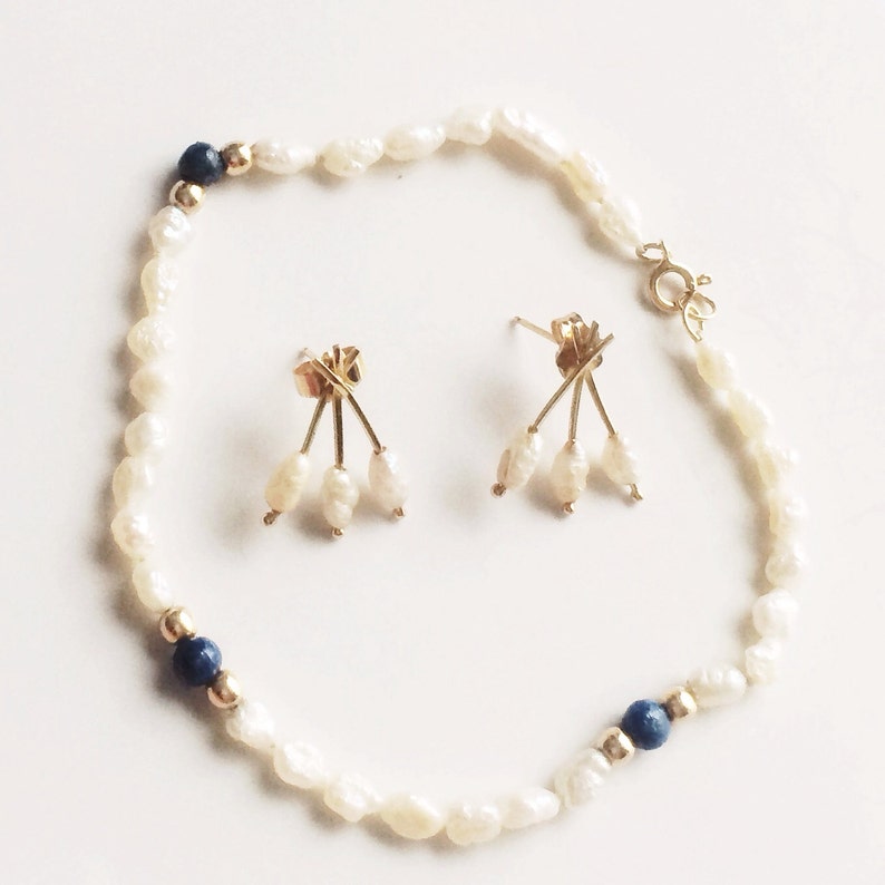 May include: A set of pearl earrings and a pearl bracelet with blue accents. The earrings feature three pearls dangling from a gold setting. The bracelet is made of white pearls and has two blue stones.