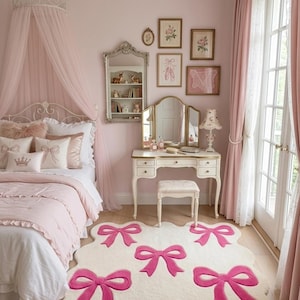 Può includere: Un tappeto color crema con bordo smerlato caratterizzato da cinque grandi motivi a fiocco rosa acceso. Il tappeto è posizionato su un pavimento in legno chiaro in una camera da letto con pareti rosa, letto a baldacchino, toeletta e sgabello.