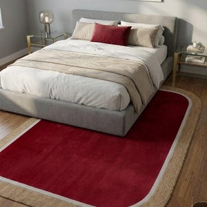 May include: A rectangular area rug with rounded corners featuring a deep red center, a thin white border, and a wide outer trim made of natural tan woven jute fiber. The rug is positioned on a wooden floor partially underneath a gray upholstered bed.