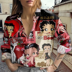Peut inclure: Une chemise boutonnée à motifs avec un collage d'illustrations de Betty Boop en rouge, blanc et noir. La chemise présente diverses images de Betty Boop, des cœurs et des cerises. Le texte sur la chemise comprend "Betty Boop", "A Doop" et "Just like the moon, I go through phases."