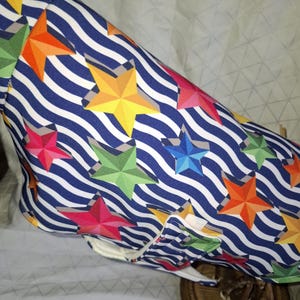 May include: A fabric item with a blue and white wavy striped pattern, adorned with colorful, geometric stars in shades of red, orange, yellow, green, and blue. The item has a white interior and a pocket with green star accents.
