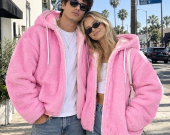Puede incluir: Dos personas con chaquetas a juego de color rosa brillante, de manga larga, con capucha y cremallera, hechas de un material suave y peludo. Ambas chaquetas tienen cordones blancos en la capucha y un cierre de cremallera blanco al frente.