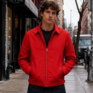Könnte beinhalten: Eine leuchtend rote Jacke mit Reißverschluss, klassischem Kragen, langen Ärmeln und zwei Vordertaschen mit silberfarbenen Druckknöpfen. Die Jacke wird über einem schlichten schwarzen T-Shirt und einer dunklen Hose getragen und wirkt schlicht.