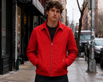 Puede incluir: Una chaqueta roja brillante con cremallera, cuello clásico, mangas largas y dos bolsillos delanteros con botones de presión plateados. La chaqueta se lleva sobre una camiseta negra lisa y pantalones oscuros, mostrando un diseño limpio.