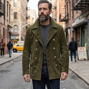 Puede incluir: Chaqueta verde oliva de doble botonadura con botones dorados. La chaqueta tiene un cuello con solapas y dos bolsillos delanteros. La persona lleva una camiseta gris de cuello redondo y vaqueros azul oscuro. El fondo muestra una calle de la ciudad.