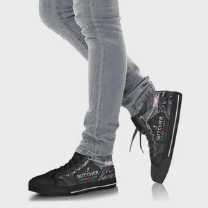 May include: High-top canvas sneakers featuring a dark gray and black patterned design with The Witcher Wild Hunt logo printed on the side. The shoes have black rubber toe caps, black laces, and a thick black rubber sole with a white stripe detail.