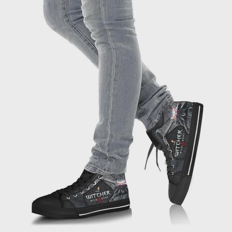 May include: High-top canvas sneakers featuring a dark gray and black patterned design with The Witcher Wild Hunt logo printed on the side. The shoes have black rubber toe caps, black laces, and a thick black rubber sole with a white stripe detail.